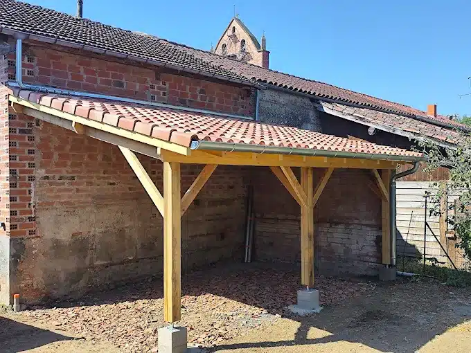 Abri de jardin neuf avec charpente bois et tuiles canal rouges, adossé à un mur de briques anciennes sous un ciel bleu.