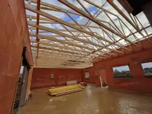 Charpente bois neuve installée dans une construction en parpaings rouges, sol boueux, chantier ouvert au ciel bleu.