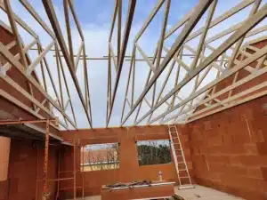 Charpente de ferme en bois posée sur des murs en briques rouges (terre cuite) lors d'une construction, avec un ciel nuageux visible.