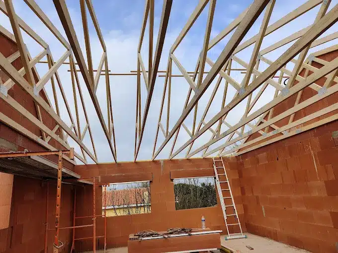 Charpente de ferme en bois posée sur des murs en briques rouges (terre cuite) lors d'une construction, avec un ciel nuageux visible.