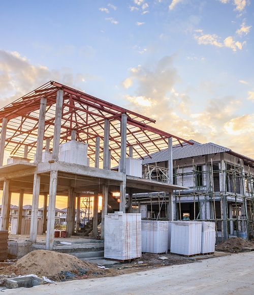 Construction de deux maisons modernes avec charpente métallique, toiture et échafaudage, sous un ciel de crépuscule. Blocs de béton sur le chantier résidentiel.