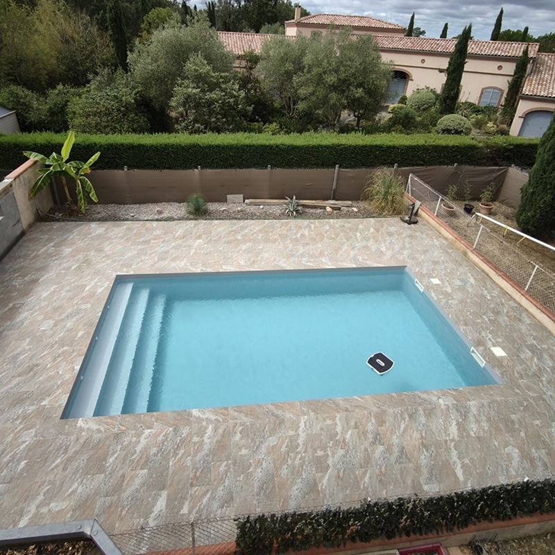 Piscine rectangulaire bleue avec nettoyeur robot, entourée d'une terrasse en carrelage beige. Vues sur maisons et verdure.