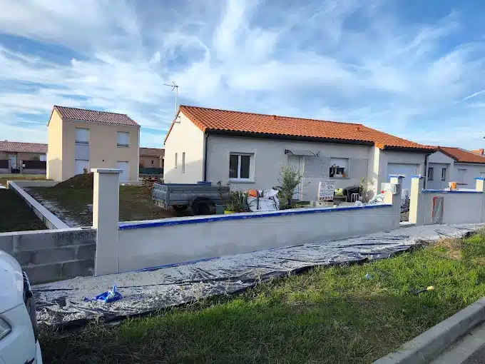 Construction Maison Neuve : Aménagement Extérieur & Clôture Lotissement avec maisons aux toits tuiles rouges, dont une en construction avancée. Un mur de clôture est en finition, avec matériaux et gravats au sol. Ciel bleu.