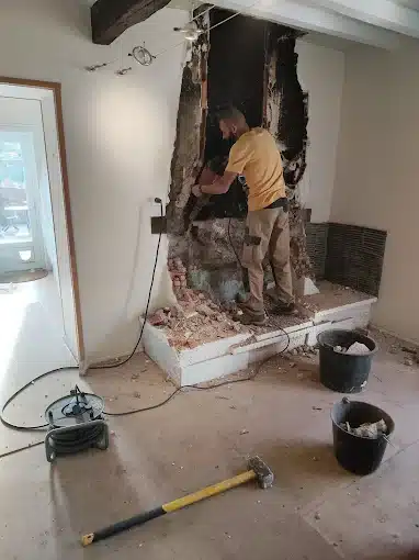 Homme barbu démolissant une ancienne cheminée en brique avec un marteau-piqueur. Débris au sol et outils de rénovation.