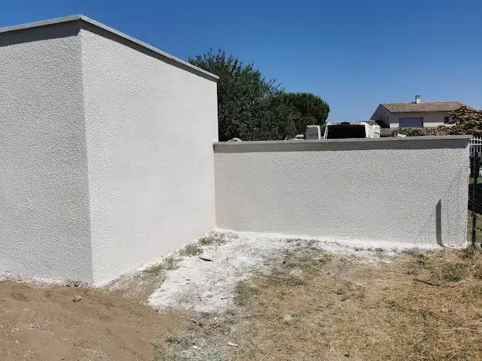 Façade et mur extérieur enduits d'un crépi clair texturé, sur un sol de chantier, sous un ciel bleu dégagé. Rénovation de bâtiment.