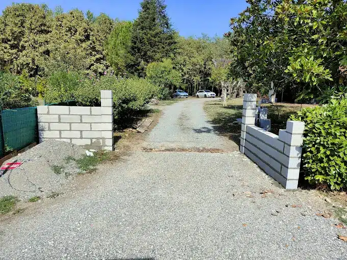 Entrée de propriété en gravier avec murs bas en parpaings blancs en construction. Deux voitures visibles au loin sur l'allée entourée de verdure.