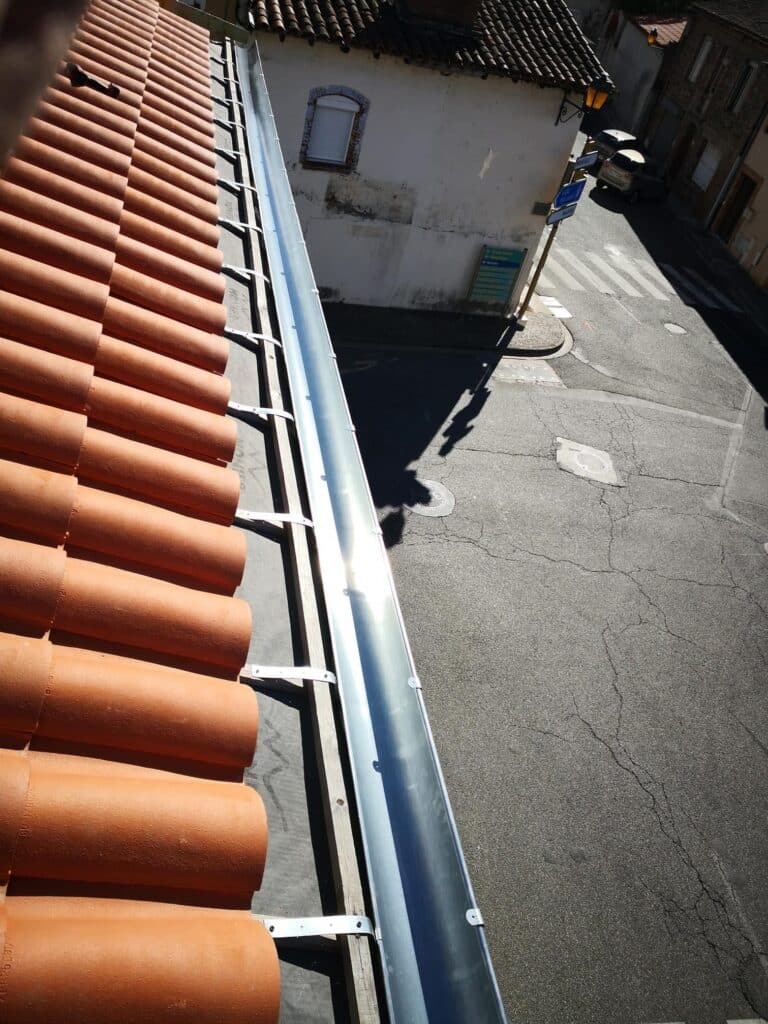Vue plongeante sur une gouttière métallique neuve installée sous des tuiles canal rouges et sur une rue asphaltée avec marquage au sol et panneau "Notaire".