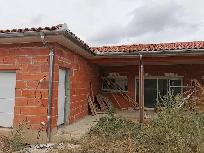Maison neuve en briques rouges, toit tuiles TERREAL et gouttières. Porte garage, porte d'entrée et menuiseries installées. Chantier extérieur.