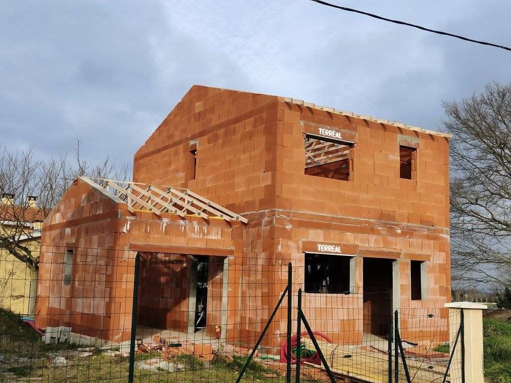 Construction de maison individuelle en parpaings rouges TERREAL, charpente partielle visible, clôture provisoire au premier plan, ciel nuageux.
