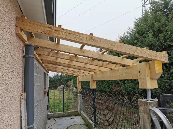 Construction d'une pergola en bois adossée à une maison, avec charpente brute et poteaux de soutien.
