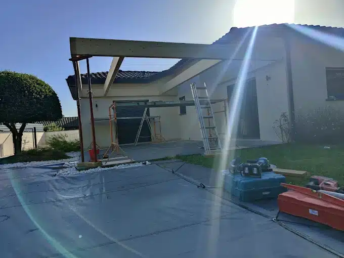 Construction d'une pergola en bois devant une maison par temps ensoleillé. Poutres supportées par des étais, échelle et outils de chantier sur bâche.