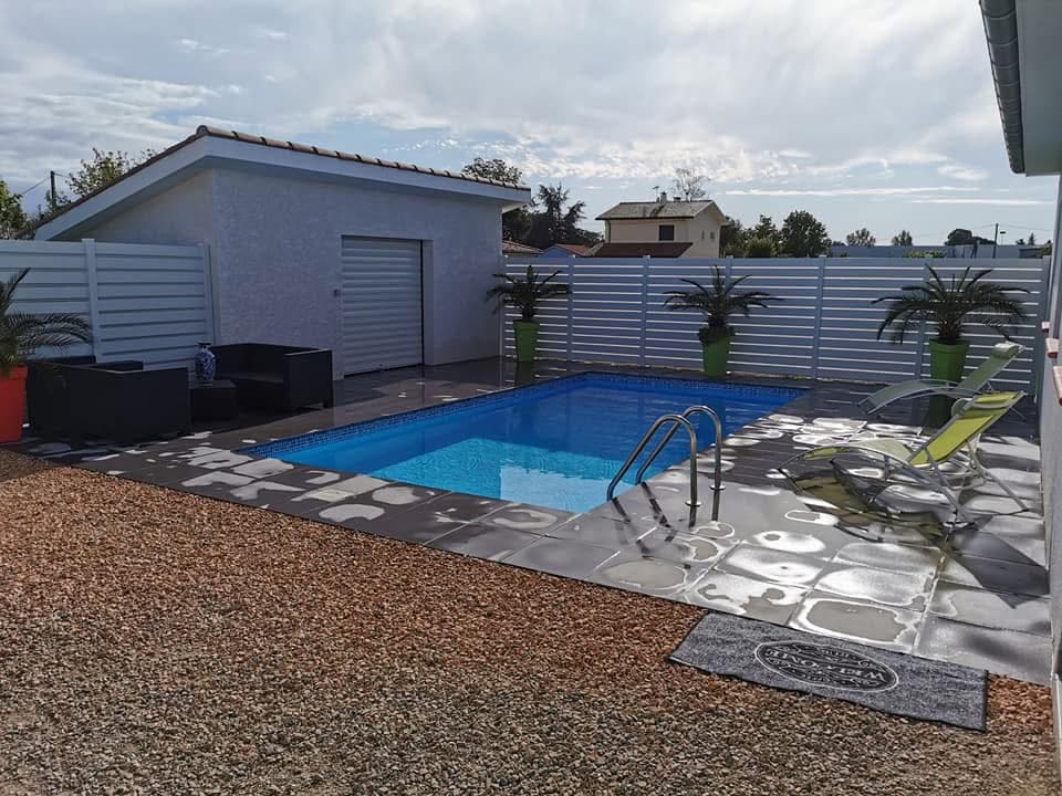 Piscine rectangulaire avec margelle sombre et gravier brun, clôture blanche et cabanon. Tapis "WELCOME".