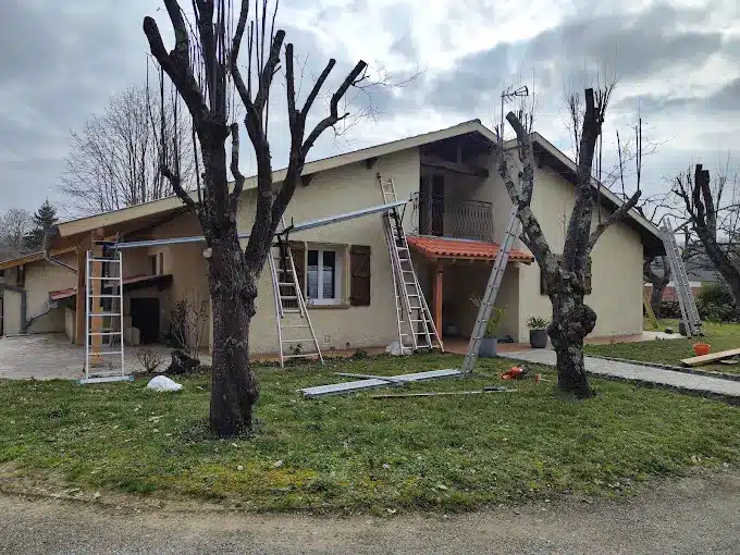 Maison beige en rénovation avec des échelles appuyées et des éléments de gouttière en cours d'installation, entourée d'arbres taillés et d'un jardin.