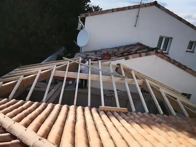 Rénovation de toiture avec charpente bois en cours de pose, un ouvrier travaille sur le toit d'une maison aux tuiles anciennes.